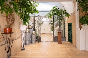 A corridor with a tiled floor, potted plants, a large window, and a door with a green shutter.