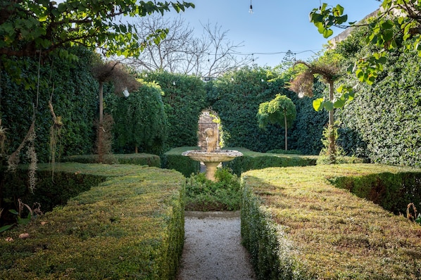 A formal garden with a central fountain, trimmed hedges, and a gravel path.