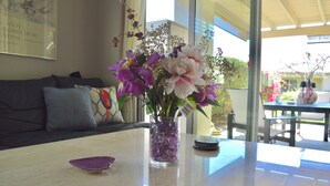 A living room with a glass vase of flowers on a table, a sofa with cushions, and a window with a view of the outdoors. - Resort Style Home- Pool Table- Walking Distance to Coachella Fest & Stagecoach (Indio)