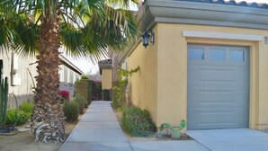 A residential street with a single-story house featuring a garage door, a palm tree, and a well-maintained garden. - Resort Style Home- Pool Table- Walking Distance to Coachella Fest & Stagecoach (Indio)