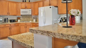 A kitchen with wooden cabinets, a white refrigerator, a microwave, and a granite countertop. - Resort Style Home- Pool Table- Walking Distance to Coachella Fest & Stagecoach (Indio)