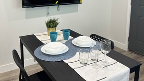 Comedor con una mesa negra, mantel blanco, individuales azules y un televisor de pantalla plana montado en la pared.