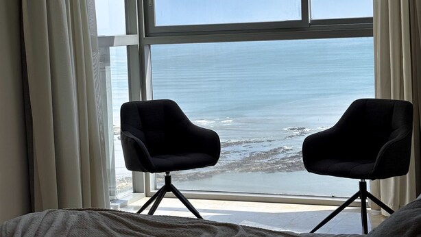 A hotel room with a large window overlooking the ocean, two black chairs, and a bed with a grey blanket. - Departamento de lujo Yoo-Panamá 34B  (Panama City)