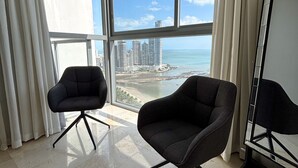 A spacious room with two black armchairs, a large window with a city and beach view, and a reflective floor. - Departamento de lujo Yoo-Panamá 34B  (Panama City)