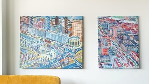 A room with a mustard-colored sofa, two colorful cityscape paintings on the wall, and a vintage record player on a wooden stand. - Central Panorama Apartment | Chmielna 35 (Warszawa)