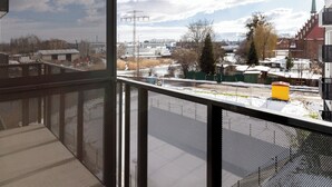 A view from a window with a metal mesh screen, overlooking a snowy landscape with buildings, a church spire, and a clear sky. - Mostek Sapphire Navy Studio (Gdansk)