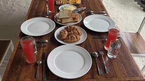 A wooden dining table set with plates, cutlery, and food items including bread, croissants, and a fruit plate. - Mandhoo Inn (Mandhoo)