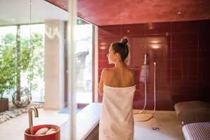 Un espace de détente avec un mur de verre, une personne enveloppée dans une serviette rouge, un seau rouge rempli de pierres et un lit recouvert d’une couverture à carreaux.