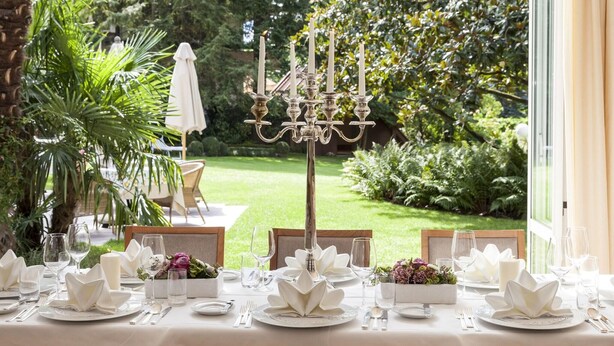 Une table dressée pour un repas élégant en plein air, avec vue sur un jardin bien entretenu et agrémentée d’un chandelier en guise de centre de table.
