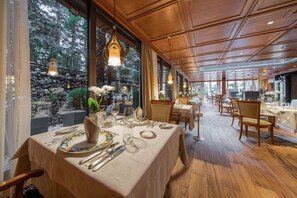 A restaurant with wooden flooring, tables set with white tablecloths, and hanging pendant lights. - Mignon (Merano)