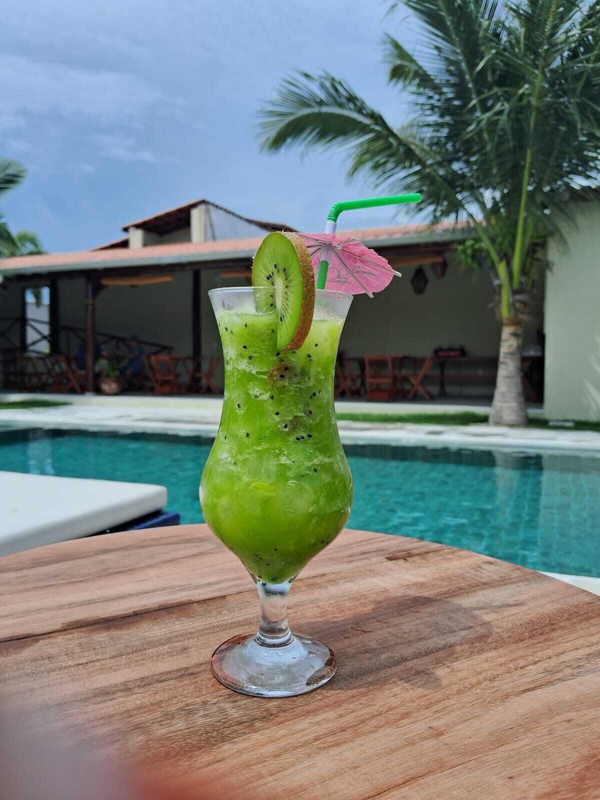 A green cocktail with a kiwi slice and a straw, placed on a wooden table by a pool.