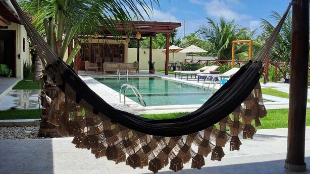 A hammock hanging over a swimming pool with a view of a building and palm trees. - Pousada Villa Recanto do Mar  (Amontada)