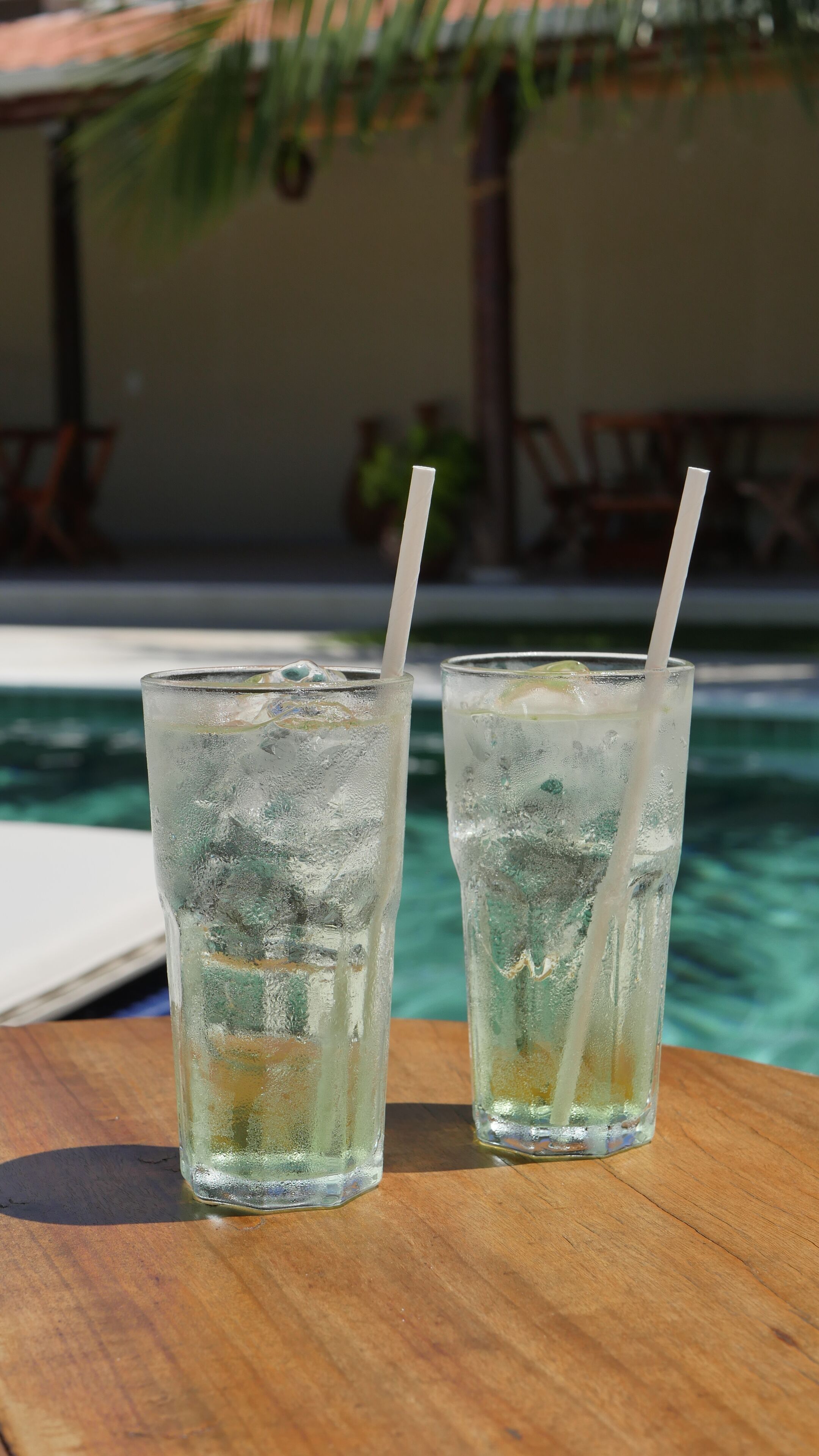 Two glasses of iced beverage with straws on a wooden table by a poolside.