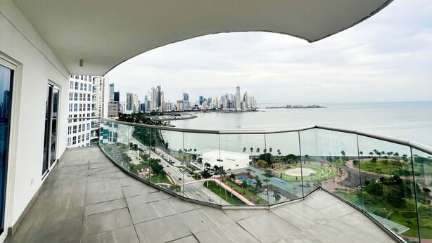 Un balcón con barandilla de vidrio curvo que da a una vista panorámica de la ciudad y al agua.