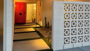 A modern building entrance with a red door, a concrete wall with geometric cutouts, and a pathway leading to an interior space.