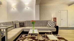 A modern living room with a sectional sofa, a coffee table, and a patterned rug. There are wall-mounted lights and a door with a number sign.