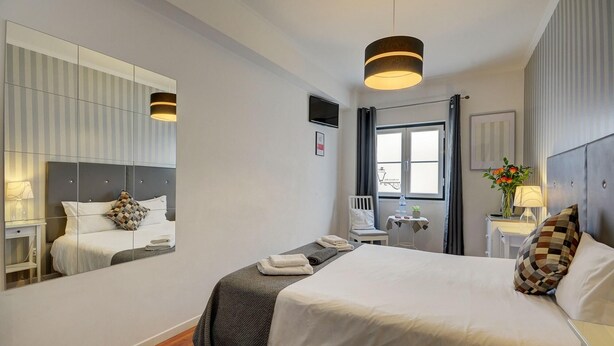 Une chambre d’hôtel moderne avec un grand lit, une table de chevet avec une lampe, une petite table avec une plante, un miroir et une fenêtre avec des rideaux.