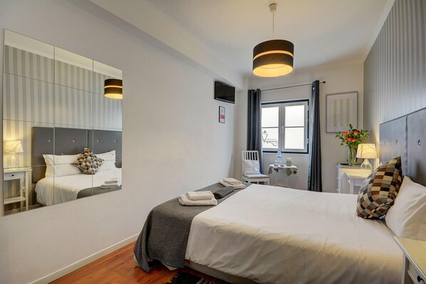 Una habitación de hotel moderna con una cama grande, una mesita de noche con lámpara, una pequeña mesa con una planta, un espejo y una ventana con cortinas.