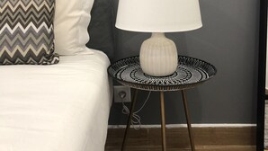 Un lit bien fait, avec une tête de lit matelassée grise, un oreiller blanc et un autre oreiller à motifs. Une lampe blanche, dont le pied est à motifs noir et blanc, est posée sur une petite table ronde à côté du lit.