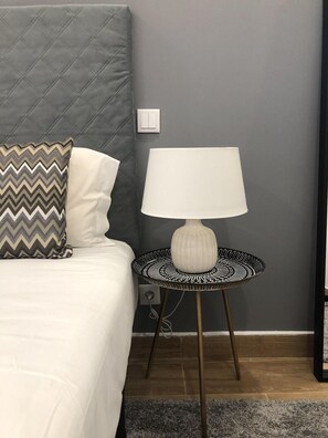 Una cama bien hecha con un cabecero gris acolchado, una almohada blanca y una almohada estampada. Una lámpara blanca con base de estampado blanco y negro está sobre una mesita redonda al lado de la cama.