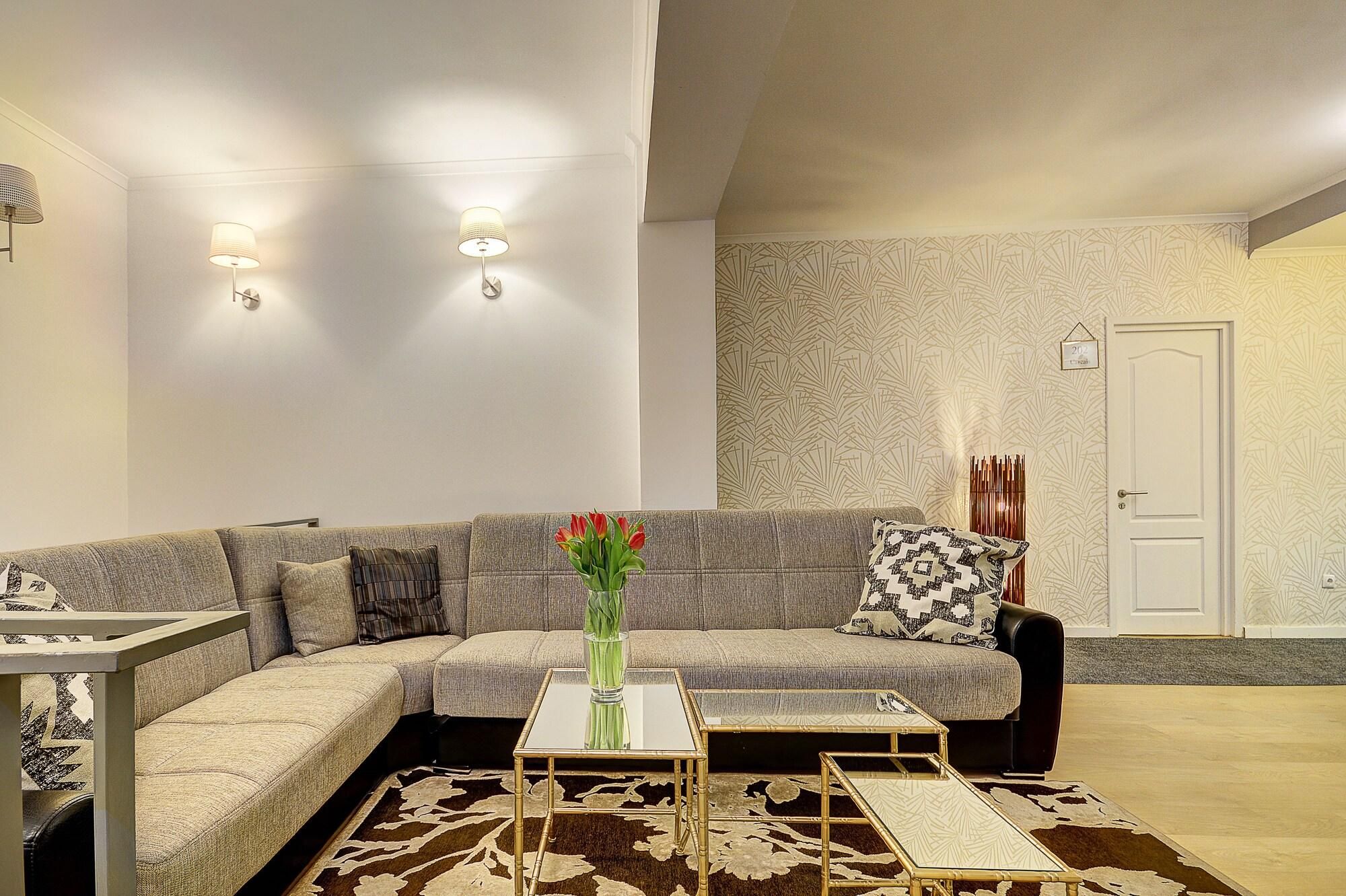 A modern living room with a sectional sofa, a coffee table, and a patterned rug. There are wall-mounted lights and a door with a number sign.