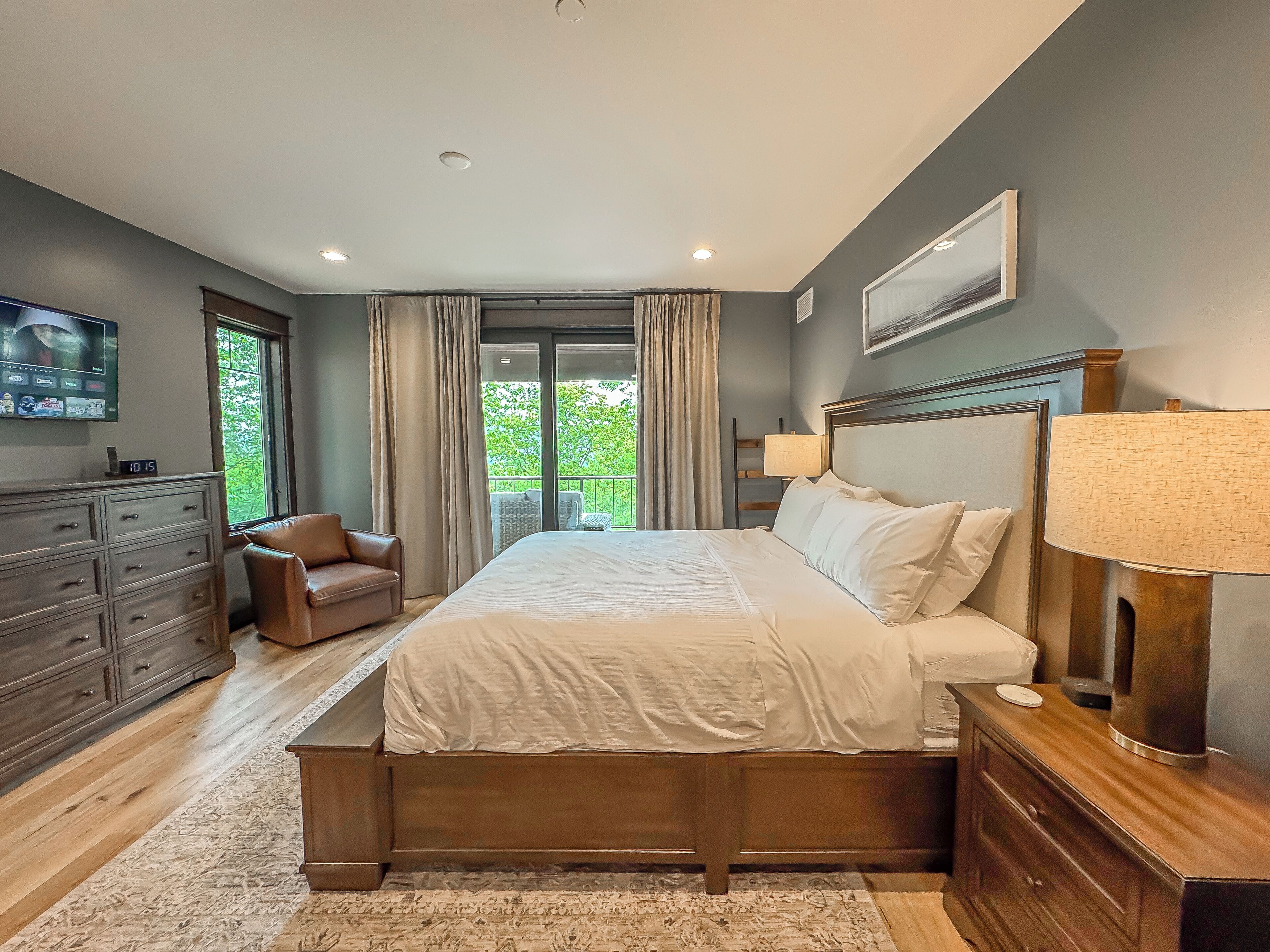 A bedroom with a large bed, a wooden dresser, a chair, a TV, and a balcony with curtains.