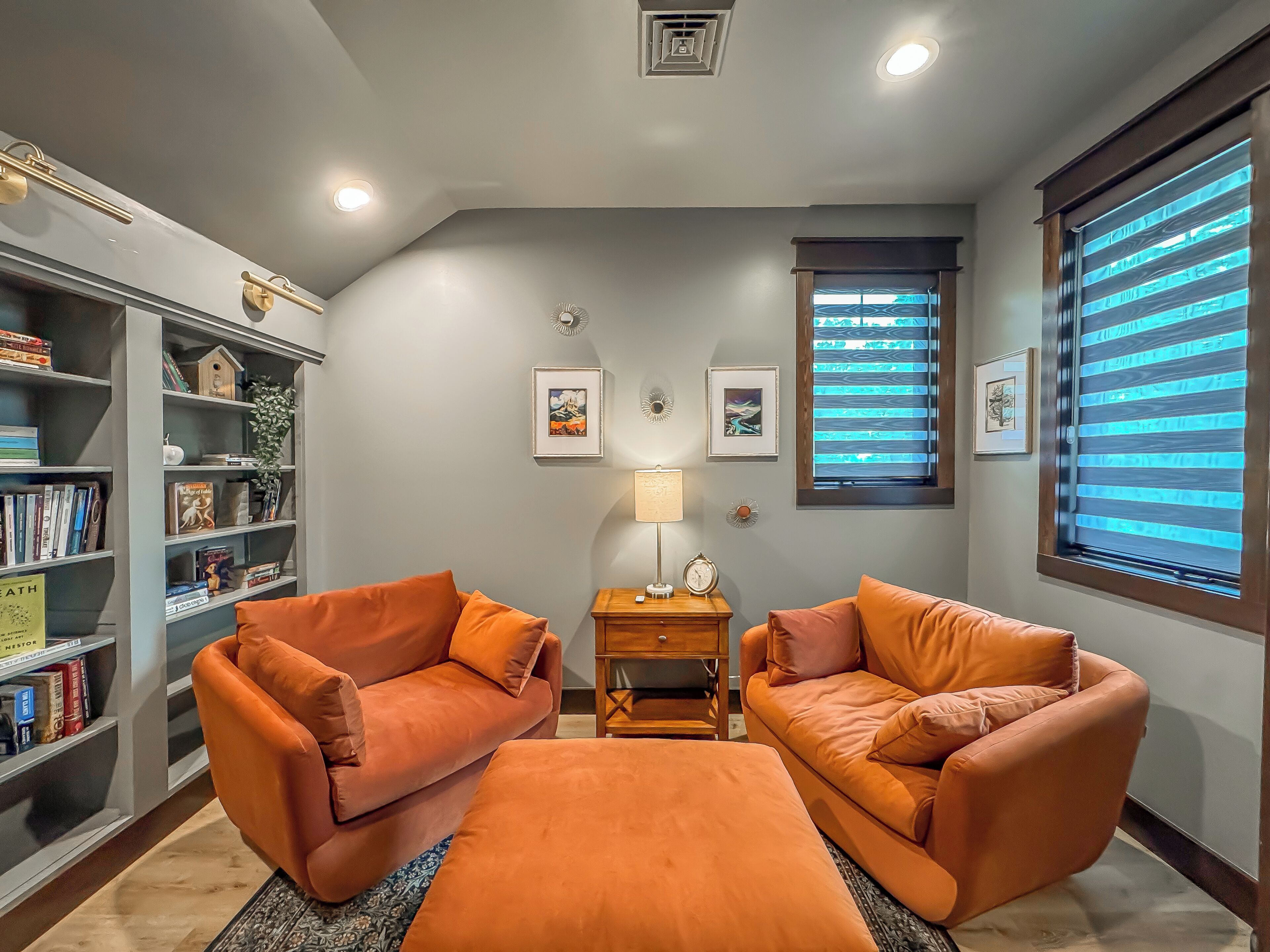 Ein gemütliches Wohnzimmer mit zwei orangefarbenen Sofas, einem Couchtisch, einem Bücherregal und einem Fenster mit Jalousien.