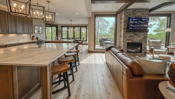 A modern kitchen with a large island, bar stools, and pendant lighting. There is a stone fireplace and a seating area with a view of the outdoors. - New + Stunning + Luxury! Lake Access Home + Community Pool! (McHenry)