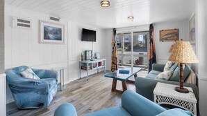 A room with a wooden ceiling, a television mounted on the wall, a sofa, a chair, a small table, and a lamp. - The Iron Seahorse by Carolina Getaway - Rooftop Deck & Stunning Ocean Views (Surf City)