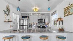 A modern kitchen with a central island, bar stools, and decorative elements like a mermaid tail and anchor wall art. - The Iron Seahorse by Carolina Getaway - Rooftop Deck & Stunning Ocean Views (Surf City)