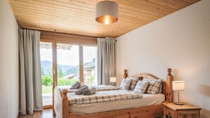 A wooden bed with a headboard, two bedside tables, and a view of mountains through a large window. - Le Souvenir by Interhome (Ovronnaz)