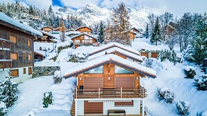 A snow-covered mountain lodge with multiple wooden cabins, a central building with a balcony, and a view of snow-covered trees and mountains. - Le Souvenir by Interhome (Ovronnaz)
