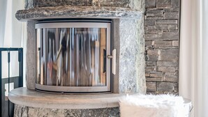 A stone fireplace with a glass door, a white fluffy stool, and a stone wall in the background. - Le Souvenir by Interhome (Ovronnaz)