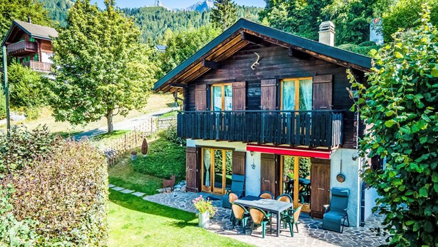 A two-story wooden house with a balcony, surrounded by lush greenery and mountains in the background. - Le Coeur by Interhome (Ovronnaz)