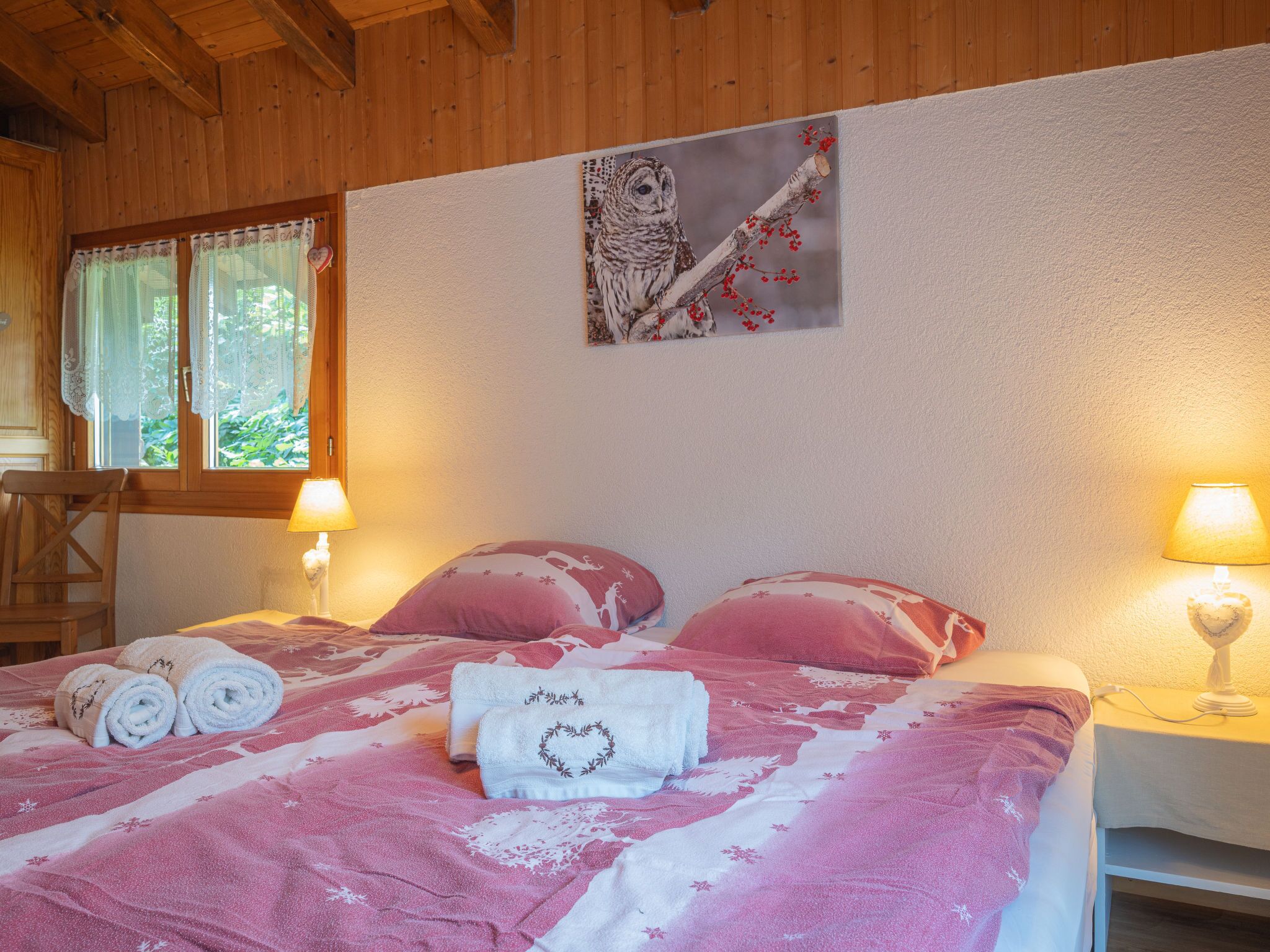A cozy bedroom with a bed, two bedside lamps, a window with lace curtains, and a painting of an owl on the wall.