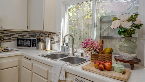 Una cocina con armarios blancos, un microondas, un tostador, un lavamanos y una ventana con vistas a una valla de madera y árboles.