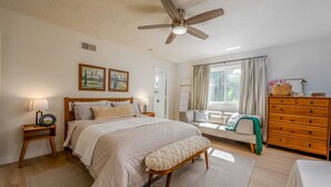 Un dormitorio con cama, banco, cómoda, mesita de noche, ventilador de techo y ventana con cortinas.