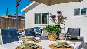 Un área de comedor al aire libre con una mesa lista para cuatro personas, rodeada de sillas, una planta en maceta y un parasol.