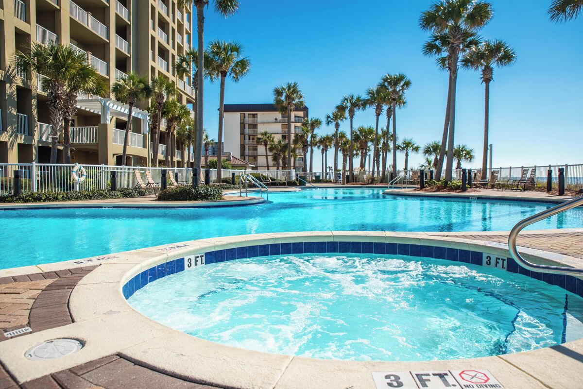 A swimming pool with a hot tub, surrounded by palm trees and a multi-story building.