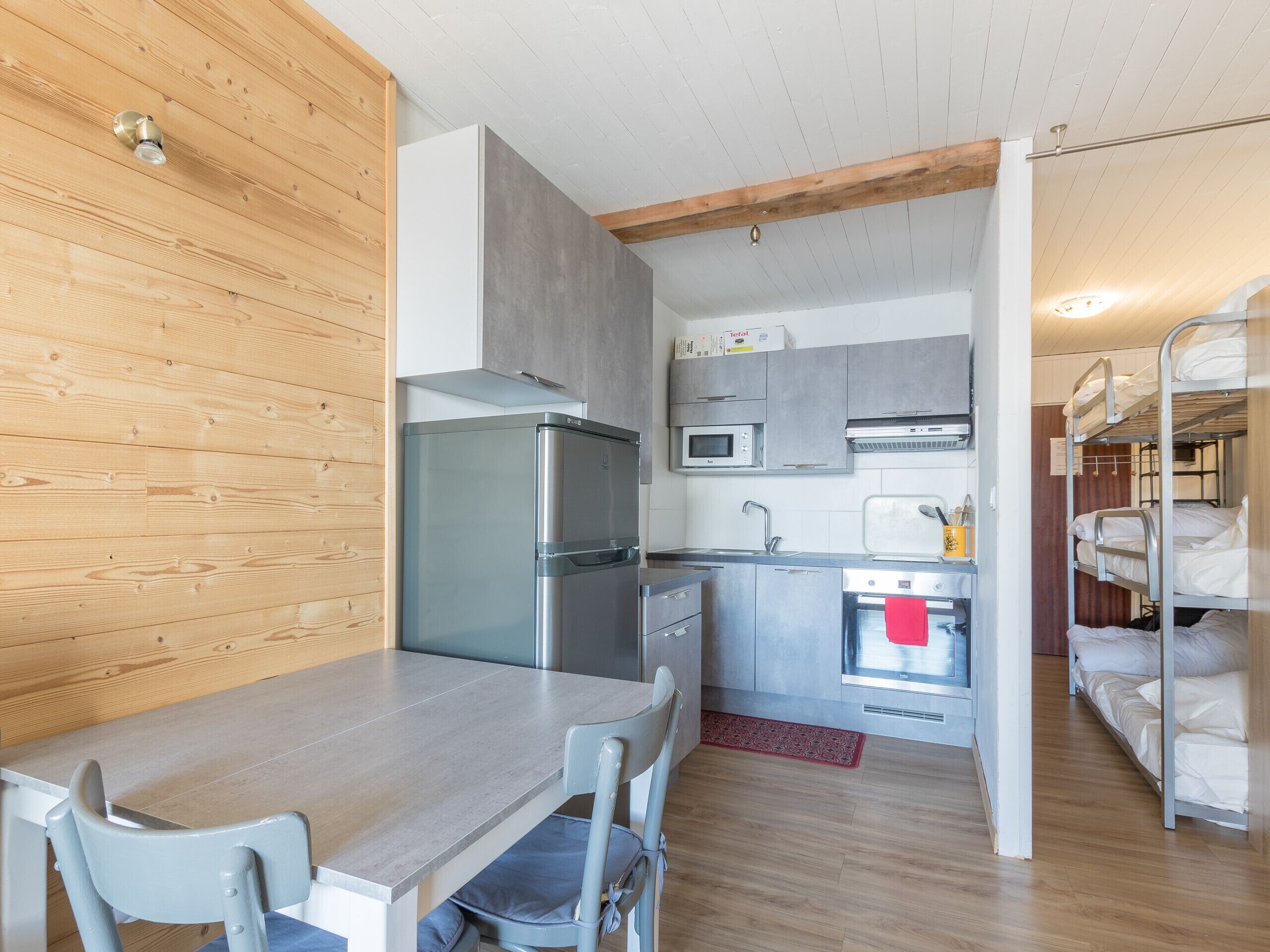 Une cuisine compacte équipée d’un réfrigérateur, d’un four à micro-ondes, d’un four et d’un évier. On y trouve une table à manger avec des chaises et un lit superposé avec du linge de lit blanc.
