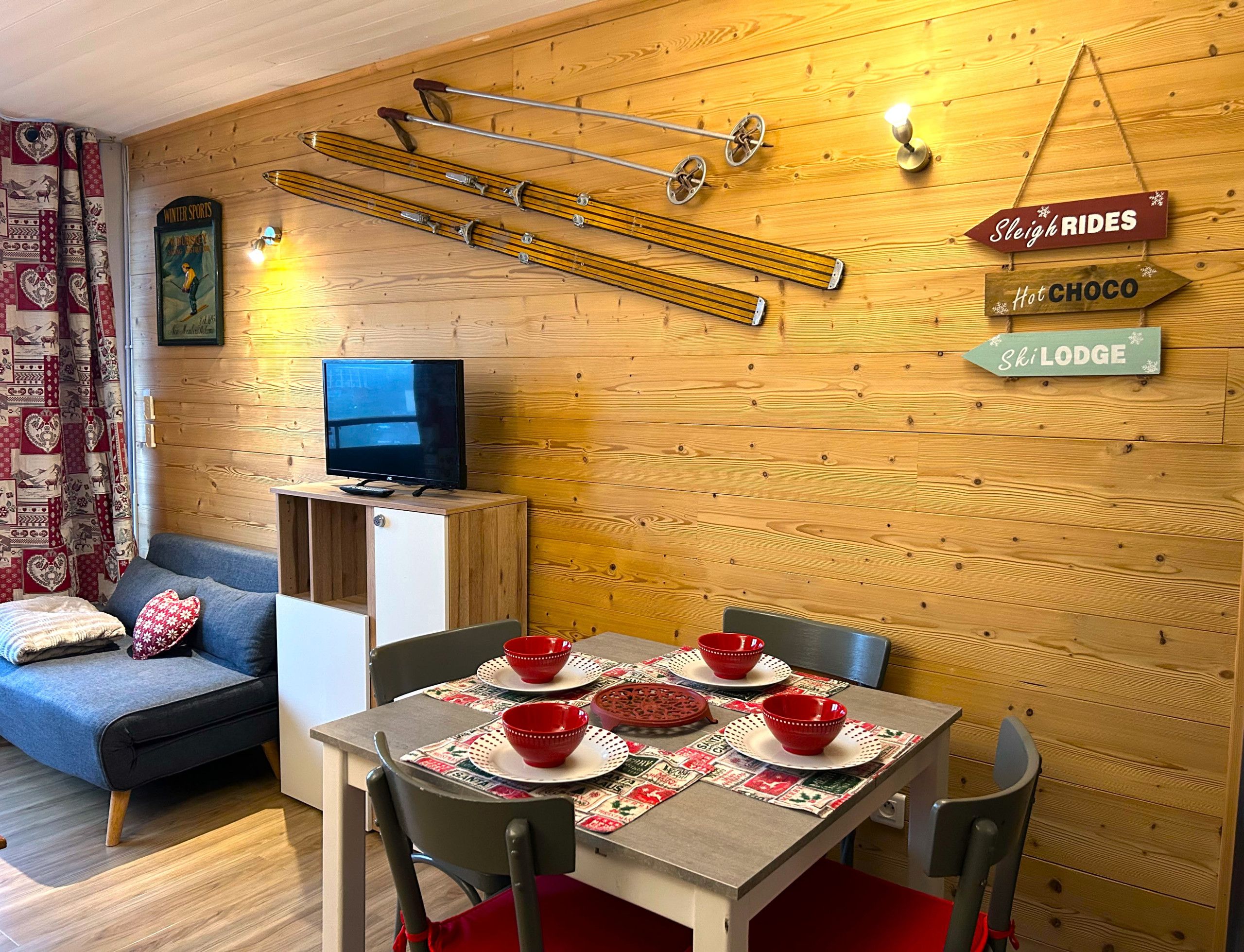 A cozy room with a wooden wall, a television on a cabinet, a dining table with red bowls, and a sign indicating 'Sleigh Rides' and 'Hot Choco'.