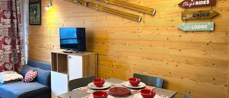A cozy room with a wooden wall, a television on a cabinet, a dining table with red bowls, and a sign indicating 'Sleigh Rides' and 'Hot Choco'.