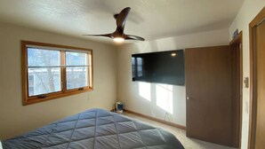 Un dormitorio con cama, ventilador de techo, ventana, televisor montado en la pared y puerta.