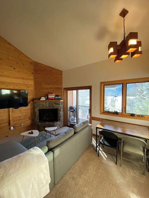 A cozy living room with a stone fireplace, wooden walls, a flat-screen TV, a sofa, a dining table with chairs, and a large window offering a view of the outdoors.