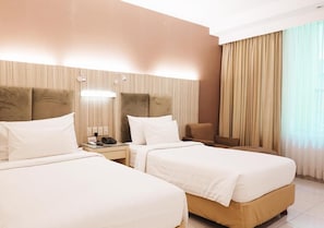 A hotel room with two beds, a bedside table with a lamp, a chair, and a large window with curtains. - Hotel Wisata Niaga (Purwokerto)