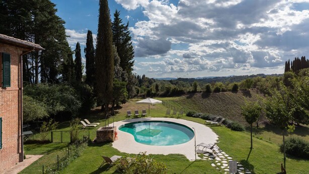 Une piscine entourée d’une pelouse, avec un bâtiment en briques et de grands arbres en arrière-plan.
