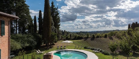 Una piscina rodeada de césped, con un edificio de ladrillo y altos árboles al fondo.