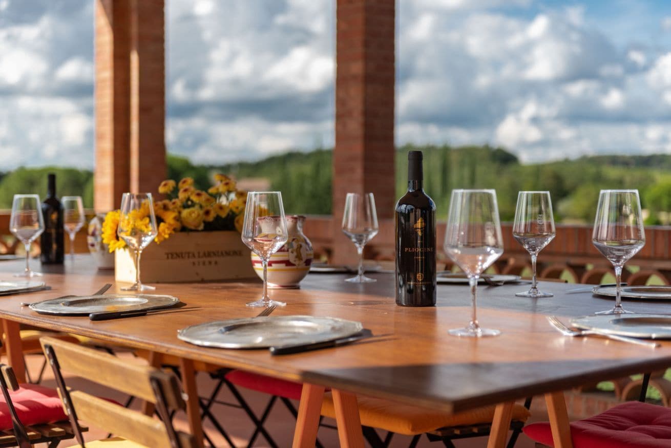 A dining table set for a meal with wine glasses, plates, and cutlery, with a bottle of wine and a vase of flowers.