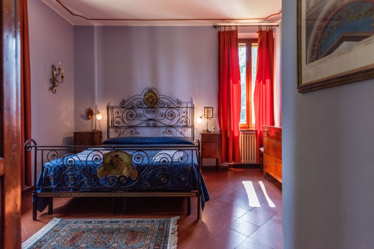 Un dormitorio con estructura de cama metálica, colcha azul, alfombra estampada, cómoda de madera y ventana con cortinas rojas.