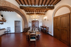 Un comedor con una larga mesa de madera dispuesta para una comida, rodeada de sillas. Hay una araña colgando del techo, y las paredes están decoradas con arcos de ladrillo y puertas de madera.